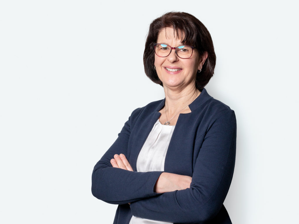 Professionelle Frau mit Brille, die lächelnd mit verschränkten Armen vor einem hellen Hintergrund steht. Sie trägt eine elegante Bluse und einen blauen Blazer, was auf einen geschäftlichen Kontext hindeutet.