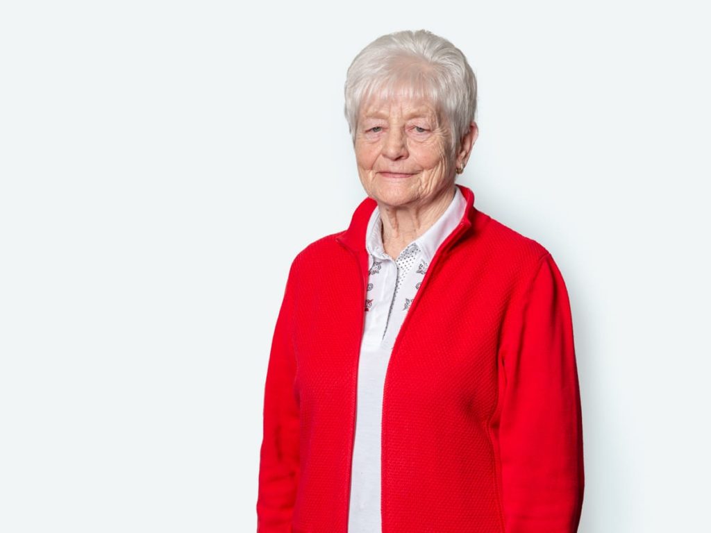 Ältere Frau mit grauen Haaren in einem roten Pullover, die lächelnd vor einem hellen Hintergrund steht.