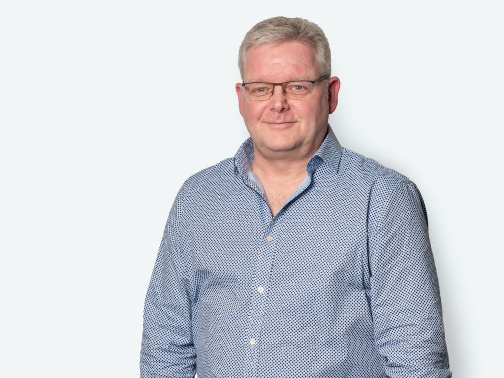 Mann mit Brille und blauer, gepunkteter Hemd steht vor einem hellen Hintergrund und lächelt freundlich.
