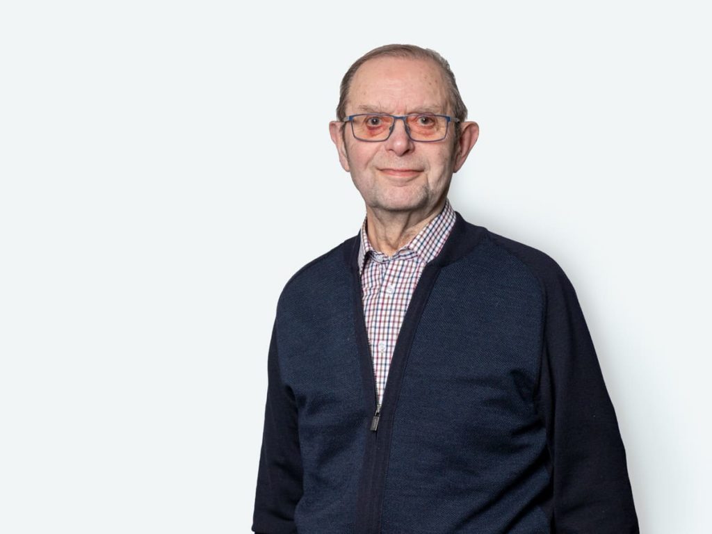 Älterer Mann mit Brille und grauen Haaren, der in einem dunkelblauen Pullover vor einem hellen Hintergrund steht. Er lächelt freundlich und trägt ein kariertes Hemd.