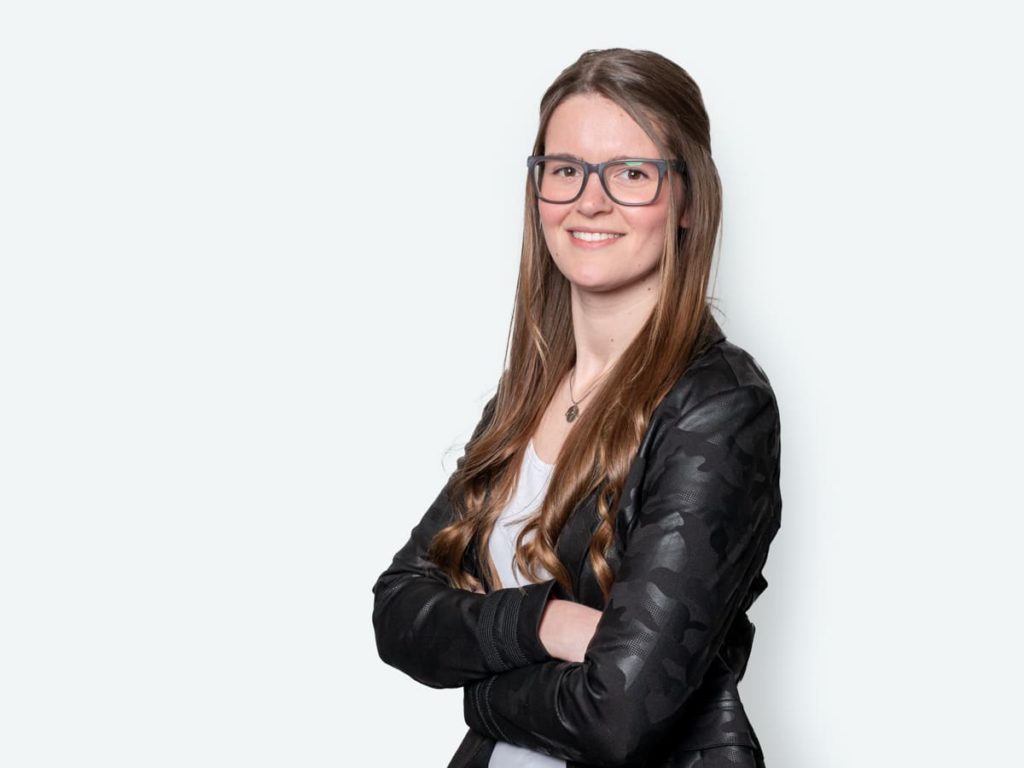 Junge Frau mit langen, lockigen Haaren und einer schwarzen Jacke, die in die Kamera lächelt und die Arme verschränkt. Hintergrund in sanften Pastellfarben.