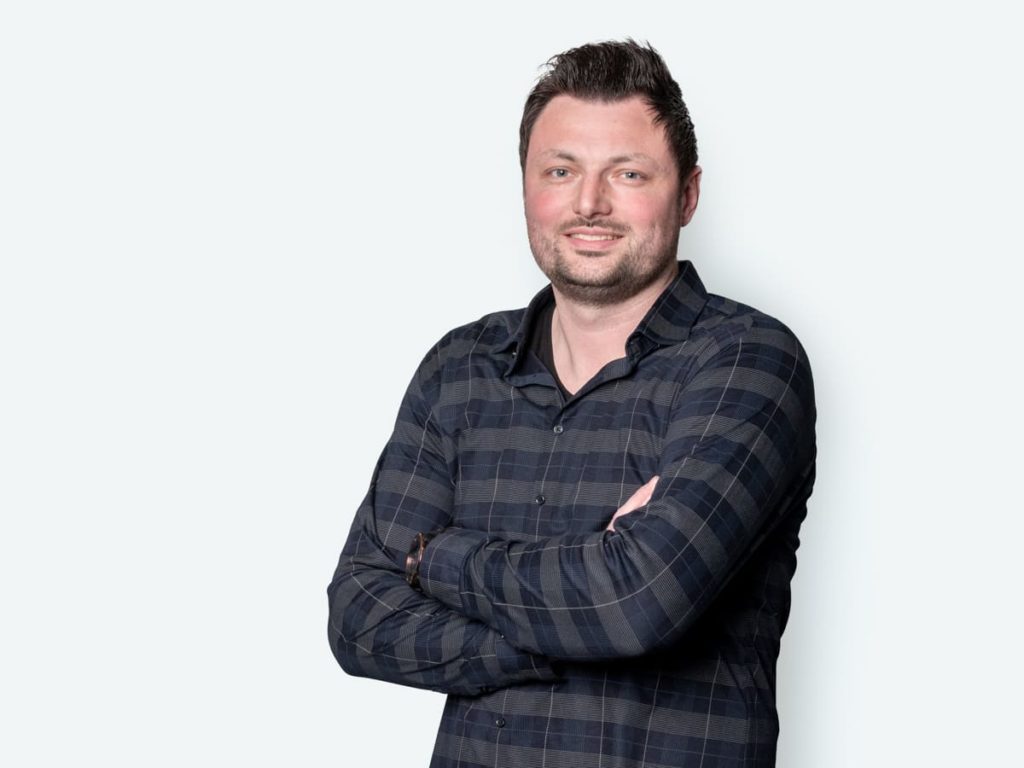 Mann mit verschränkten Armen, der in einem karierten Hemd vor einem hellen Hintergrund steht, lächelt selbstbewusst.