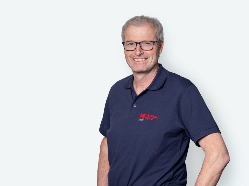 Mann mit Brille und grauen Haaren, trägt ein dunkelblaues Polo-Shirt mit dem Logo von Metallbau Knapp, steht vor einem hellen Hintergrund und lächelt freundlich.