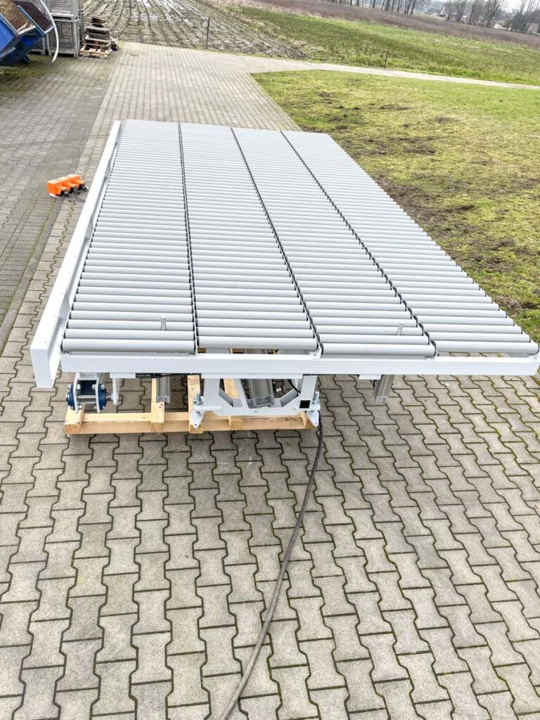 Eine große, graue Förderbandanlage auf einem Holzgestell, die auf einem gepflasterten Weg steht. Im Hintergrund sind landwirtschaftliche Felder und weitere Maschinen zu sehen. Die Anlage ist für den Transport von Materialien konzipiert.