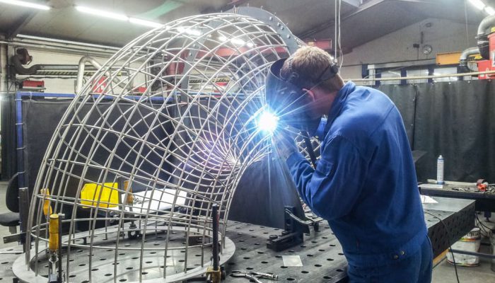 Ein Schweißer arbeitet an einem Metallrahmen in einer Werkstatt, während er mit einem Lichtbogen schweißt. Der Rahmen hat eine komplexe Gitterstruktur und wird auf einem stabilen Werkbanktisch bearbeitet. Im Hintergrund sind verschiedene Werkzeuge und Maschinen sichtbar, die für Metallbearbeitung verwendet werden.