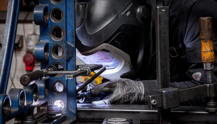 Ein Schweißer arbeitet konzentriert an einer Metallkonstruktion, während er eine Schweißmaske trägt. Im Vordergrund sind Werkzeuge und das Schweißgerät sichtbar, während der Hintergrund eine Werkstattumgebung zeigt.
