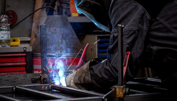 Ein Schweißer arbeitet an einem Metallrahmen, während Funken und Rauch entstehen. Die Szene zeigt eine Werkstatt mit Werkzeugen und einem Schweißgerät im Hintergrund. Der Handwerker trägt Schutzausrüstung, um sich während des Schweißens zu schützen.